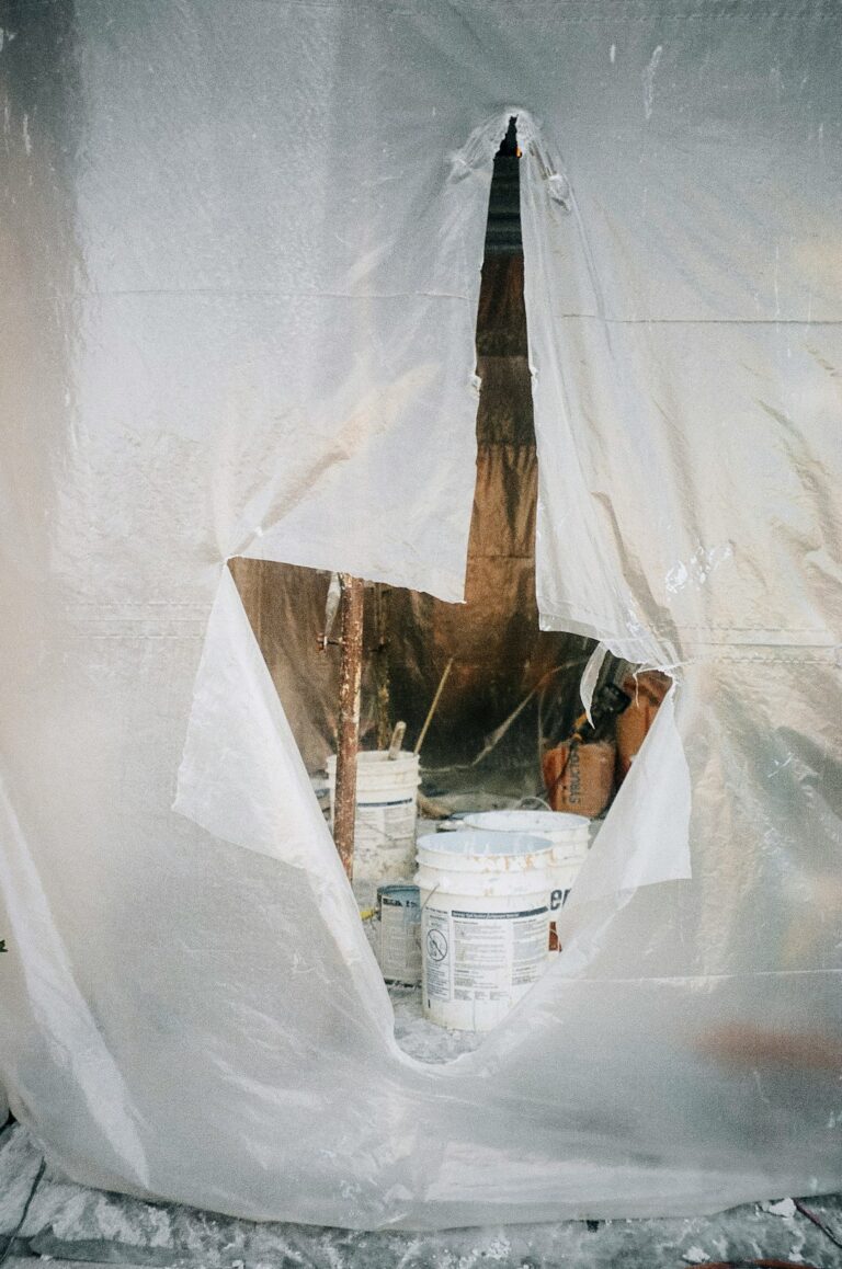 A homeowner places a bucket beneath a ceiling leak while storm-damaged shingles are visible through an attic opening.